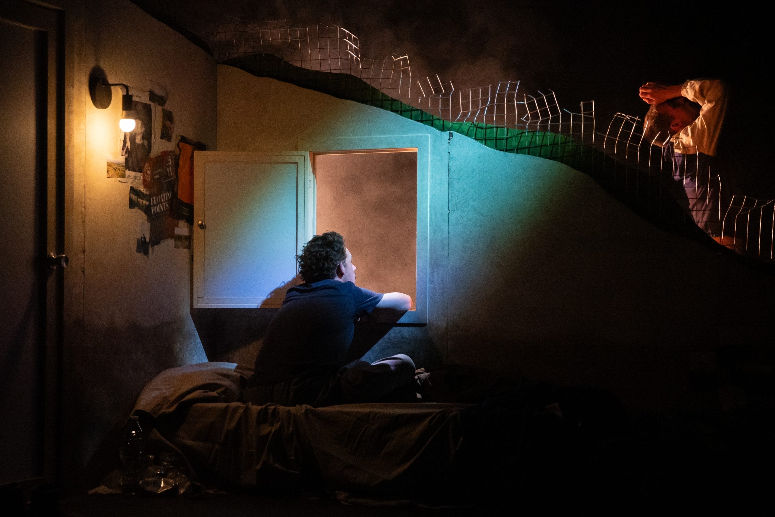 A young man sits in a dark bedroom looking out of a window.